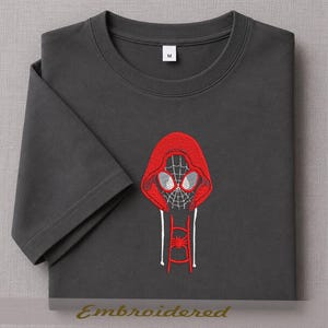 May include: Dark gray t-shirt featuring an embroidered design of a red hooded mask with a spiderweb pattern and white accents. The design includes a spider emblem on the lower portion. The shirt has a crew neck and short sleeves.
