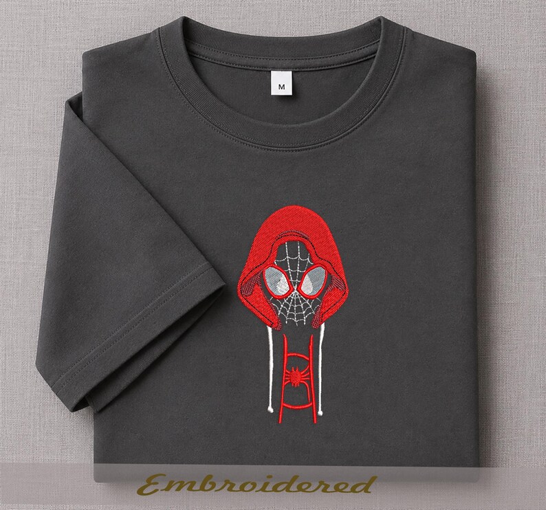 May include: Dark gray t-shirt featuring an embroidered design of a red hooded mask with a spiderweb pattern and white accents. The design includes a spider emblem on the lower portion. The shirt has a crew neck and short sleeves.