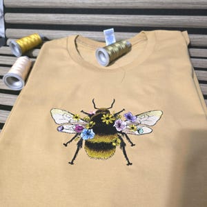 Pode incluir: Uma t-shirt amarelo-mostarda com um bordado central de um abelhão adornado com pequenos detalhes florais coloridos em tons de rosa, azul e amarelo. Três carretéis de linha de costura em amarelo e bege estão colocados nas proximidades.
