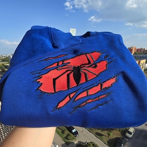 May include: A royal blue crewneck sweatshirt featuring a large embroidered design on the chest. The design depicts a red fabric patch underneath a blue layer, with a black spider emblem centered over the red area, creating a torn effect.