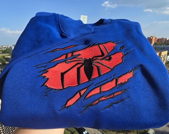 May include: A royal blue crewneck sweatshirt featuring a large embroidered design on the chest. The design depicts a red fabric patch underneath a blue layer, with a black spider emblem centred over the red area, creating a torn effect.