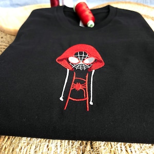 Może przedstawiać: Czarny t-shirt z wyhaftowaną grafiką postaci w czerwonej bluzie z kapturem, białym wzorem pajęczyny na masce, białymi soczewkami oczu i czerwonym emblematem pająka na klatce piersiowej oraz dwoma zwisającymi białymi sznurkami od kaptura.