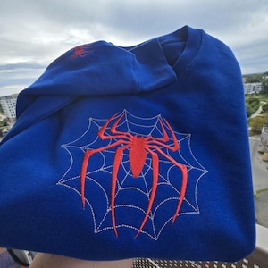 May include: A royal blue crewneck sweatshirt featuring a large, centered red embroidered spider design layered over a white stitched spiderweb pattern. A smaller, matching red spider embroidery is positioned on the upper sleeve area.