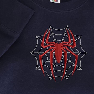 Op de afbeelding: Een donkerblauw sweatshirt met ronde hals voorzien van een grote, rode geborduurde spin in het midden van een wit gestikt spinnenwebontwerp. Een kleinere rode spin is geborduurd op de mouwboord. Het kledingstuk heeft een geribbelde halslijn.