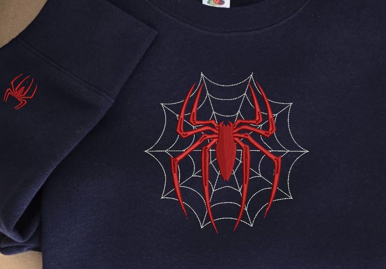 Op de afbeelding: Een donkerblauw sweatshirt met ronde hals voorzien van een grote, rode geborduurde spin in het midden van een wit gestikt spinnenwebontwerp. Een kleinere rode spin is geborduurd op de mouwboord. Het kledingstuk heeft een geribbelde halslijn.