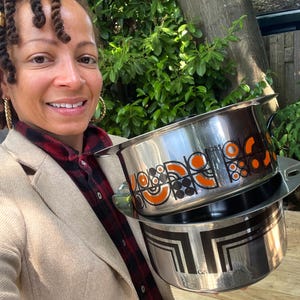 Puede incluir: Dos ollas de acero inoxidable con diseños geométricos negros y naranjas. La olla superior tiene un diseño de círculos y líneas curvas, mientras que la olla inferior presenta un patrón angular negro. Ambas ollas tienen asas y están apiladas.