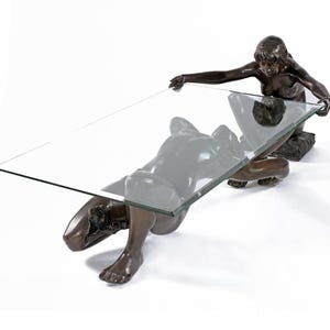 Pode incluir: Mesa de centro escultural em bronze com duas figuras sustentando um tampo de vidro retangular. As figuras estão ajoelhadas, uma parece estar deitada. A mesa tem um acabamento marrom escuro. O tampo de vidro tem cerca de 120 cm.