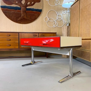 Può includere: Una scrivania moderna di metà secolo con un cassetto rosso lucido e un piano color crema. La scrivania ha una base cromata con un design angolare unico. Il cassetto ha due maniglie cromate. La scrivania è un elegante pezzo d'arredamento.