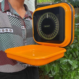 May include: A bright orange kitchen scale with a square, flat weighing platform and a vertical display panel. The display features a circular dial with black and yellow markings, numbers ranging from 0 to 1000, and the brand name EKS in the center.