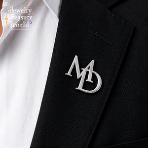 Puede incluir: Pin con monograma MD plateado sobre una blazer negra. El pin es un diseño estilizado de las letras M y D, y está sujeto a la solapa de una chaqueta de traje negra. La imagen es un primer plano.