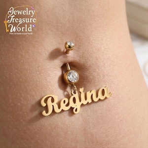 May include: Gold-colored belly button ring with a dangling nameplate that reads "Regina" in a cursive font. The ring features a clear, round gem at the bottom and a smaller gem on the top stud. The jewelry is displayed on skin.