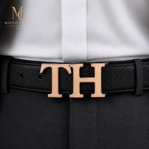 Può includere: Una cintura in pelle nera testurizzata con una grande fibbia metallica color oro rosa a forma di lettere TH. La cintura è indossata sopra pantaloni neri e una camicia bianca. Nell'angolo in alto a sinistra è presente il logo Monogram With Care.