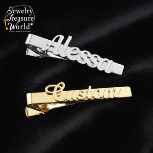 Könnte beinhalten: Zwei Metall-Krawattenklammern, eine silberne mit dem Namen "Alessa" und eine goldene mit dem Wort "Custom". Die Klammern werden auf schwarzem Hintergrund präsentiert. Das Logo von Jewelry Treasure World befindet sich in der oberen linken Ecke.