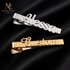 May include: Two metal tie clips with a spring-loaded clasp mechanism. One silver-toned clip features the name Alessa in a cursive script, while the other gold-toned clip features the word Custom in a cursive script, both attached to the front bars.