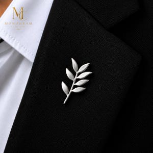 May include: A silver-toned metal lapel pin shaped like a leafy branch with six distinct leaves. The pin is attached to the black fabric of a blazer lapel, positioned next to a white shirt collar. The logo MC Monogram is visible in the top corner.