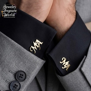 Może przedstawiać: Złote spinki do mankietów z inicjałami "MA" są prezentowane na czarnej koszuli. Osoba ma na sobie szary garnitur. Obraz pochodzi z Jewelry Treasure World.