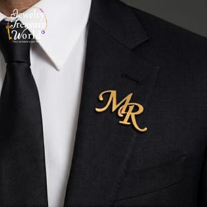 May include: A gold-colored lapel pin in the shape of the letters "MR" is pinned to a black suit jacket. The pin is a stylish accessory, perfect for formal wear. The jacket is paired with a white dress shirt and a black tie.
