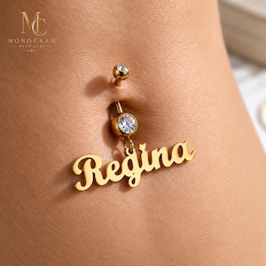 May include: A gold-toned navel piercing featuring a small round clear gemstone at the top and a larger round clear gemstone at the base. A dangling gold-toned nameplate charm with the name Regina in a cursive script font hangs from the base.
