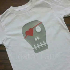May include: White t-shirt with a gray skull graphic. The skull has a red heart over one eye and a white eyepatch. The text "Alligator Lane" is printed below the skull.