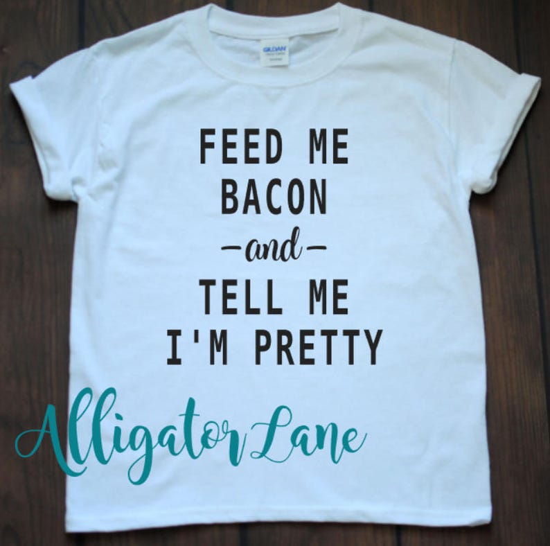 May include: White t-shirt with black text that reads "Feed me bacon -and- tell me I'm pretty". The text "Alligator Lane" is written in teal below the main text.