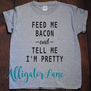 May include: Gray t-shirt with black text that reads "Feed me bacon -and- tell me I'm pretty". The text is in a casual font. The bottom of the shirt has the text "Alligator Lane" in teal.