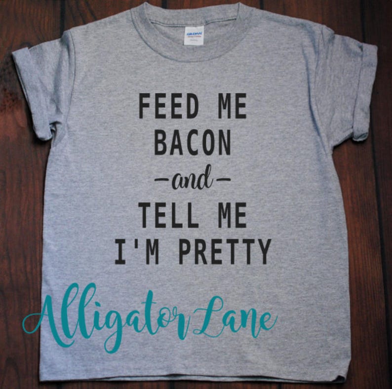 May include: Gray t-shirt with black text that reads "Feed me bacon -and- tell me I'm pretty". The text is in a casual font. The bottom of the shirt has the text "Alligator Lane" in teal.