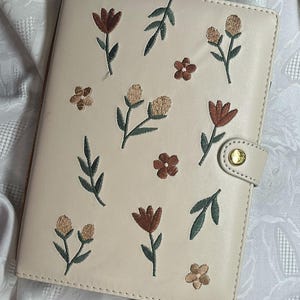 May include: Cream-colored planner with embroidered floral designs in shades of brown, tan, and green. The planner has a gold-colored snap closure. The cover is decorated with various embroidered flowers and leaves.