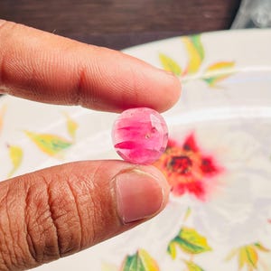 May include: A faceted, round, pink gemstone held between two fingers. The gem has a translucent quality with streaks of darker pink throughout. The background is a blurred floral design on a white surface.