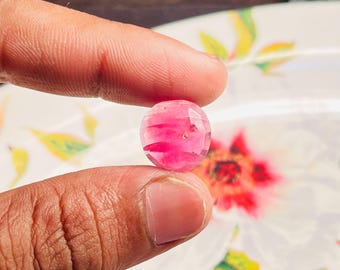 Può includere: Una gemma rosa rotonda e sfaccettata tenuta tra due dita. La gemma è traslucida con striature rosa più scuro. Lo sfondo presenta un motivo floreale sfocato su una superficie bianca.