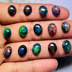 Puede incluir: Una colección de ópalos negros ovalados con destellos de color vibrantes, incluyendo rojo, verde y azul. Los ópalos se exhiben sobre una superficie clara, mostrando sus cualidades iridiscentes. Ideales para la creación de joyas.