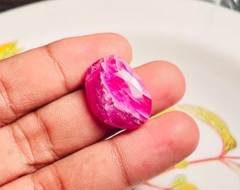 Può includere: Una gemma sfaccettata di forma ovale con una vibrante tonalità rosa e venature bianche. La gemma è tenuta in una mano, mostrando la sua superficie lucida e gli intricati motivi interni. Il colore della pietra varia dal magenta intenso al rosa pallido.