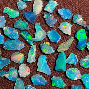 Op de afbeelding: Een verzameling ruwe opaal edelstenen in verschillende vormen en maten. De opalen vertonen een reeks kleuren, waaronder blauw, groen en flitsen van andere tinten. De edelstenen zijn verspreid over een donkerbruin oppervlak.
