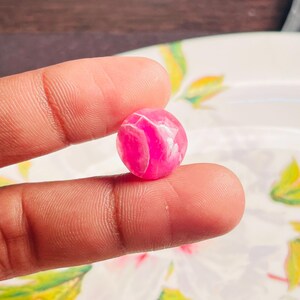 May include: A small, round, pink bead with white veining. The bead is held between two fingers, showcasing its polished surface and color variations. The bead's size is approximately 0.5 inches in diameter.
