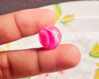 Può includere: Una piccola perla rotonda rosa con venature bianche. La perla è tenuta tra due dita, mostrando la sua superficie lucida e le variazioni di colore. La perla ha un diametro di circa 1,3 cm.