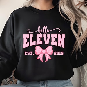 May include: Black sweatshirt with the text "hello ELEVEN" in pink, along with a pink bow graphic and the text "EST. 2015". The sweatshirt is a casual style, suitable for everyday wear.