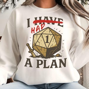 May include: White sweatshirt with a graphic featuring a large, tan, 20-sided die with the number 1, a sword, and the text "I HAVE A PLAN". The word "HAVE" is crossed out in red.