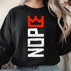 以下が含まれることがあります： 黒いスウェットシャツ。白い文字で垂直に配置された「NDP」と赤い王冠のグラフィック。クルーネックのスタイルです。