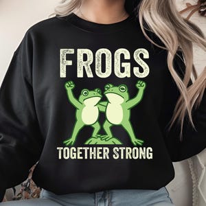 Könnte beinhalten: Schwarzes Sweatshirt mit dem Wort "FROGS" in weißer, verblichener Schrift über einer Grafik von zwei grünen Fröschen. Die Frösche umarmen sich, darunter der Text "TOGETHER STRONG".