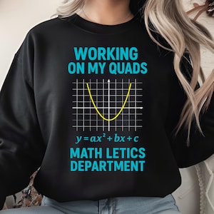 May include: Black sweatshirt with the text "WORKING ON MY QUADS" in teal above a yellow parabola graph. Below the graph is the equation "y=ax²+bx+c" and the words "MATH LETICS DEPARTMENT" in teal.