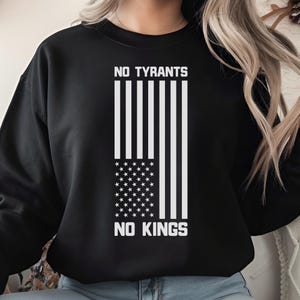 以下が含まれることがあります： 黒いスウェットシャツ。白いグラフィックは、縦型のデザインのアメリカ国旗です。「NO TYRANTS」と「NO KINGS」の文字が、それぞれ国旗の上と下にプリントされています。