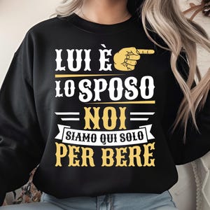 以下が含まれることがあります： 白と黄色の文字が入った黒いスウェットシャツ。「LUI È LO SPOSO - NOI SIAMO QUI SOLO PER BERE」と指を指す手のグラフィック。楽しくてカジュアルなデザインです。