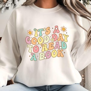 Pode incluir: Um moletom branco com a frase "IT'S A GOOD DAY TO READ A BOOK" em letras coloridas e estilo retrô. O design inclui flores e estrelas, criando uma estética alegre. É uma peça de roupa casual e confortável.