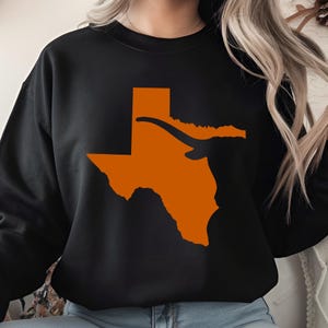 May include: A black long-sleeve crewneck sweatshirt featuring a large, solid orange graphic of the state of Texas. The design includes a stylized orange longhorn silhouette extending horizontally across the upper portion of the state shape.