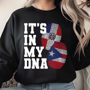 Puede incluir: Sudadera negra con el texto blanco "IT'S IN MY DNA". El diseño incluye dos formas de huellas dactilares, una con la bandera de la República Dominicana y otra con la bandera de Puerto Rico.