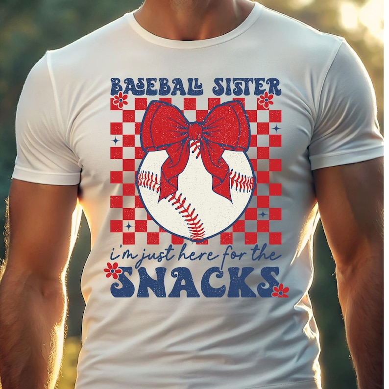 May include: White t-shirt with a baseball graphic. The design includes a baseball with a red bow, set against a red and white checkered background. Text reads "BASEBALL SISTER" and "I'm just here for the SNACKS."