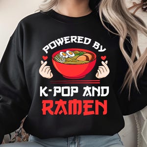 Puede incluir: Sudadera negra con el texto "POWERED BY K-POP AND RAMEN" en blanco y rojo. El diseño incluye un cuenco rojo de ramen con huevos y verduras, y dos manos haciendo formas de corazón.