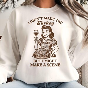 May include: White sweatshirt with a brown graphic of a woman holding a wine glass, a roasted turkey, and the text "I DIDN'T MAKE THE Turkey BUT I MIGHT MAKE A SCENE."