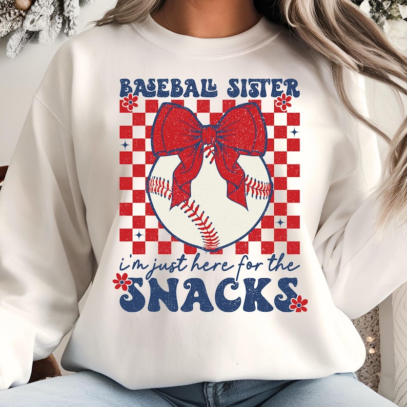 May include: White sweatshirt with a baseball-themed graphic. The design features a baseball with a red bow, set against a red and white checkered background. Text reads "Baseball Sister" and "I'm just here for the snacks."