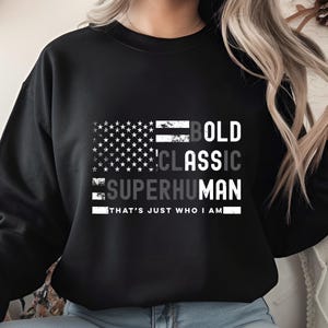Può includere: Felpa nera con un design grafico raffigurante una bandiera americana e le parole "BOLD CLASSIC SUPERHUMAN". Sotto le parole c'è la frase "THAT'S JUST WHO I AM" in bianco.