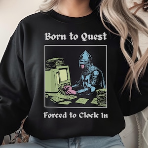May include: Black sweatshirt with the text "Born to Quest" above a graphic of a knight at a computer, and "Forced to Clock In" below. The knight is wearing armor and a helmet, and the computer is an older model.